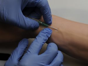 Close-up photo of latex-gloved hands inserting an IV needle into a vein in an arm.