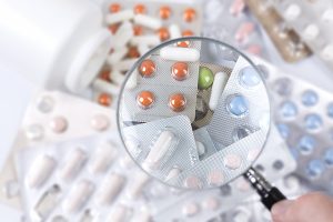 Stock photo of a variety of pills and tablets in blister packs with a magnifying glass focused on the center of the photo.